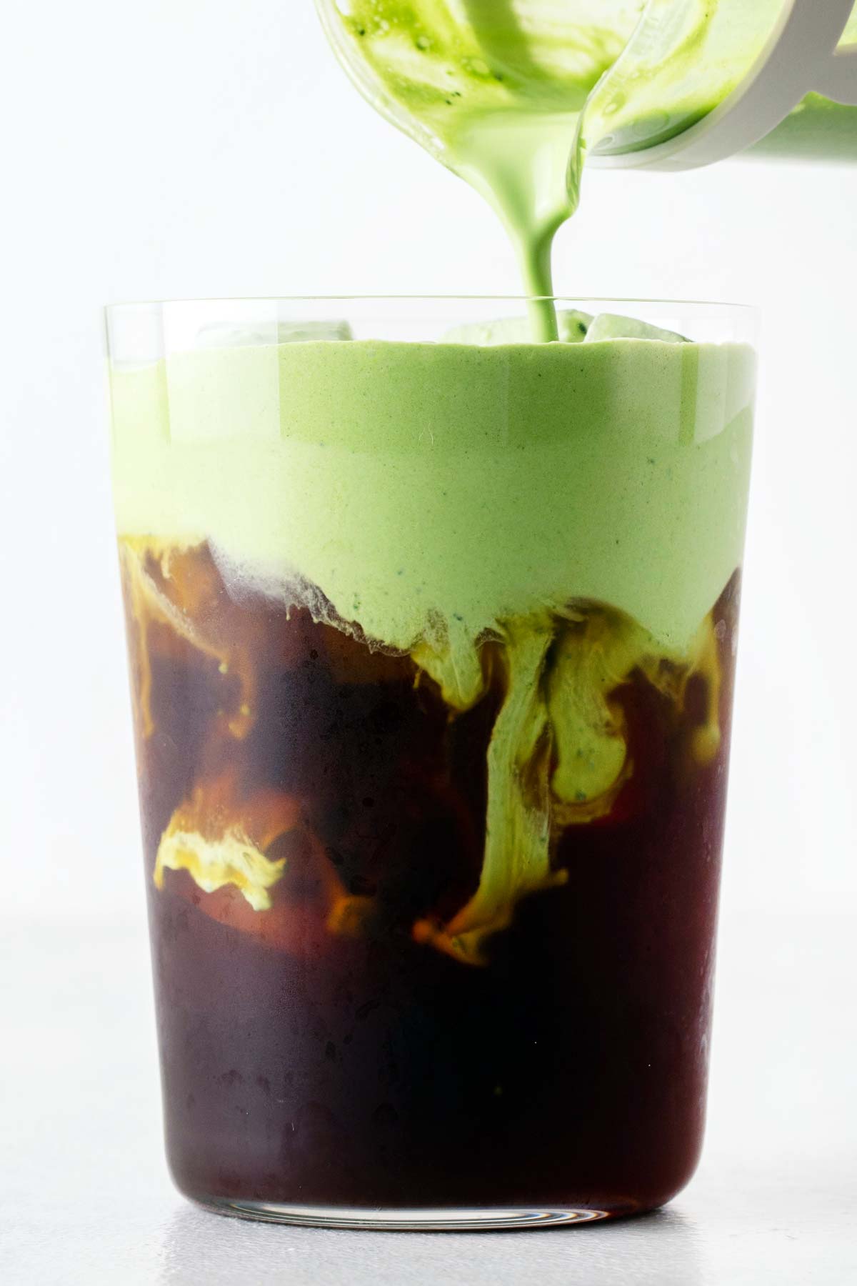 Matcha cold foam being poured over an iced coffee in a large glass.