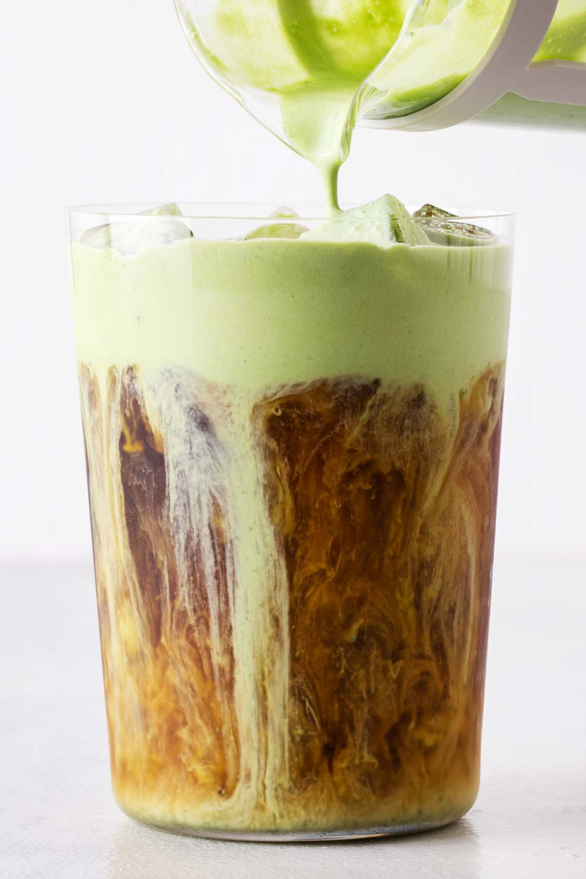 Matcha cold foam being poured over an iced coffee in a tall glass.